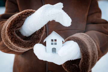 small model of a house in wool gloves on the street in winter against the background of snow