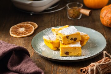 Sliced cottage cheese casserole with pumpkin and raisins, sprinkled with powdered sugar, on a ceramic plate on a brown wooden background.