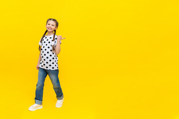 A little joyful girl with beautiful long pigtails in jeans on a yellow background.