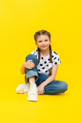 A cute little girl in blue jeans is sitting on a yellow isolated background and smiling.