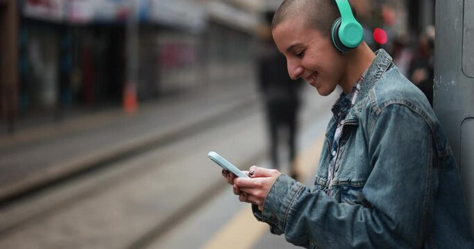 Young bald woman listening music with wireless headphones and using mobile phone while waiting at  tram station