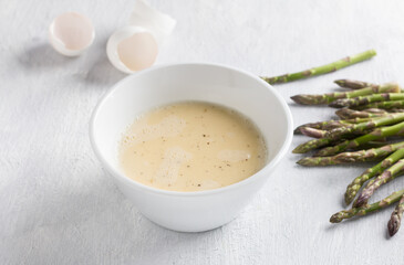 Obraz na płótnie Canvas White ceramic bowl of beaten eggs with milk and fresh asparagus shoots on a light gray background. Cooking a delicious homemade omelet