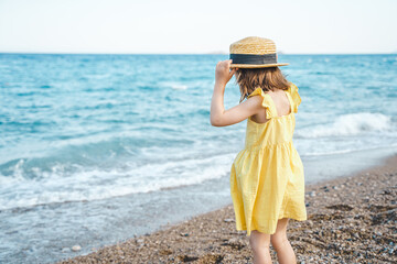 Little girl walking on beach, sea ocean shore in romantic yellow dress, straw hat. Playing in sand,...