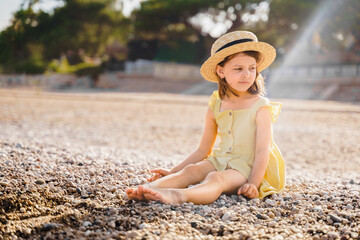 Obraz premium Little girl walking on beach, sea ocean shore in romantic yellow dress, straw hat. Playing in sand, blue waves. Family vacation travel leisure in hot summer coast. Sunny day relax in hotel resort