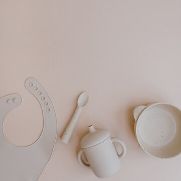 Set of silicone bowl, spoon, bib, mug for little baby kid to eat food and drink. Light pastel pink background. Flat lay, top view