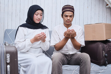 Muslim couple sitting in sofa with suitcase and luggage, palms raises praying dua before going mudik to hometown at Eid Mubarak celebration.