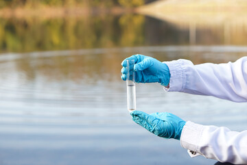 Closeup ecologist hands wears blue gloves holds test glass tube that contain sample water at the lake. Concept, explore, inspect quality of water from natural source.                      - obrazy, fototapety, plakaty