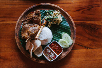 A wooden plate of deep-fried duck with rice, vegetables, chili, and crackers. Bebek Goreng. Flat lay or top view. Indonesian dish.