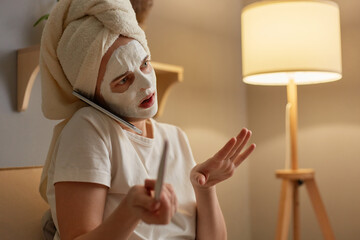Indoor shot of shocked surprised astonished woman wearing white T-shirt and in towel sitting on cough near lamp, doing manicure and talking on phone with friend, hearing breaking news.