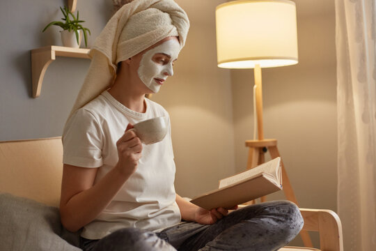 Indoor shot of young adult woman with clay mask on face wearing white T-shirt and in towel sitting on cough near lamp, holding and reading book, enjoying novel, drinking hot coffee or tea.