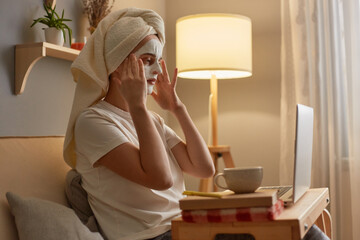 Portrait of attractive woman wearing white T-shirt and towel sitting on sofa at home, doing skin care procedures, watching film on laptop, enjoying weekend.