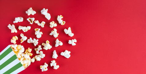 Tasty cheese popcorn falling out of a green striped carton bucket, isolated on red background. Scattering of popcorn grains. Movies, cinema, fast food and entertainment concept. Top view, flat lay