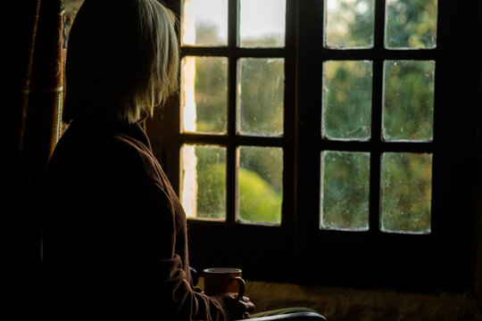 A Woman Sits Near The Window With A Cup Of Coffee. Loneliness, Sadness Concept. Midlife Crisis, Reflections.