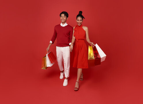 Happy asian couple holding hands going shopping center with shopping bags isolated on red background.