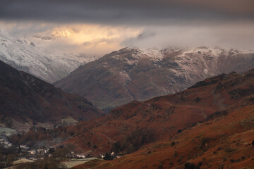 Fototapeta na wymiar Dramatic view of Lake District snowcapped mountains shrouded in low cloud on a Winter morning with small Cumbrian village; Chapel Stile.