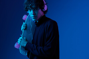 Teenage male with headphones listening to music and dancing and singing with skateboard in hand over his head, hipster lifestyle, blue background, neon light, style and trends, mixed light, copy space