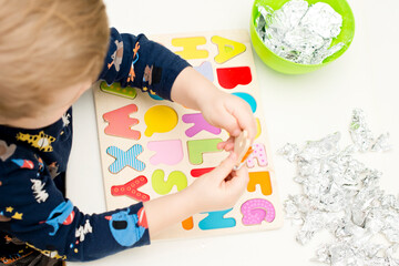 Two year boy playing with wooden alphabet letters board. Letters wrapped in foil. Intellectual game, preschool implement for early education. Verbal and memory training exercise.
