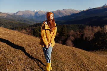 Fototapeta premium Woman smile with teeth full-length walk happiness walking on the hill and looking at mountains in a yellow raincoat and jeans in the autumn happy journey in the sun sunset hiking, freedom lifestyle 