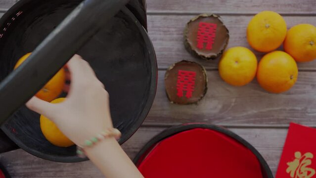 An Asian woman is putting oranges and Chinese New Year cakes into a tiffin or basket called Bakul Siah with red envelopes attached. Top view, the background is a wooden table.
