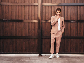 Naklejka premium Handsome confident stylish hipster model.Sexy modern man dressed in elegant beige suit. Fashion male posing in the street background in Europe city at sunset. Near wooden fence
