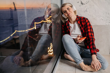 Obraz na płótnie Canvas Blonde American girl in casual plaid shirt and blue jeans sitting on windowsill using headphones, phone listening music enjoys sunset and city lights being in romantic moos. Dreamer, woman in love.
