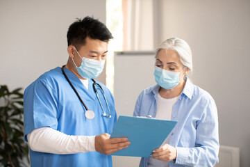 Serious middle aged korean doctor in protective mask giving contract sign to old caucasian female patient