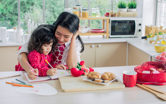 Beautiful Asian mother and mixed race adorable little daughter sitting in kitchen, drawing, painting, doing homework or activities and making card on Valentine day together, smiling with happiness.