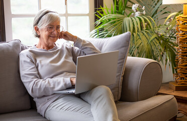 Happy 70s white-haired woman sitting on couch in living room, using laptop watching movie. Older...