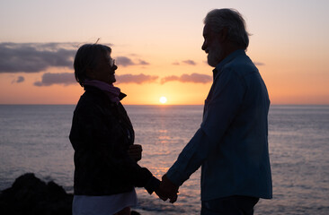 Silhouette of senior couple standing on the rocks at sea looking each other in the eyes while the sun goes down. Relaxed caucasian man and woman enjoying vacation or retirement