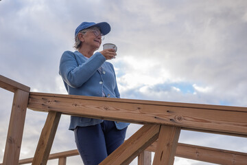 Relaxed handsome senior woman with cap and eyeglasses standing outdoor at sunrise holding a cup of coffee or tea. Smiling elderly lady enjoying silence of the morning on the beach