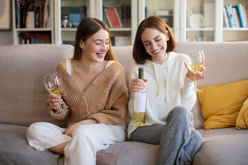 Laughing european young girlfriends with glasses and bottle of wine talking, enjoy spare time, sitting on sofa