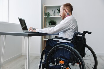 A man in a wheelchair looks at the camera businessman in the office working on a laptop online,...