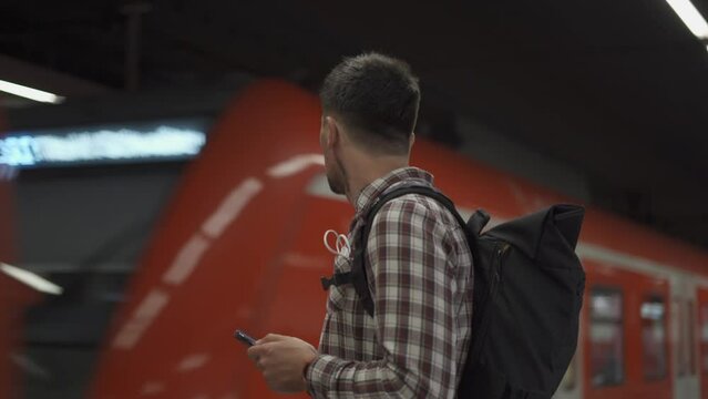 Man with smartphone in front of passing train in Munich, Germany. Passenger with backpack uses phone to create route maps and buy tickets online. S-Bahn Munchen. Muenchen municipal commuter railway. 