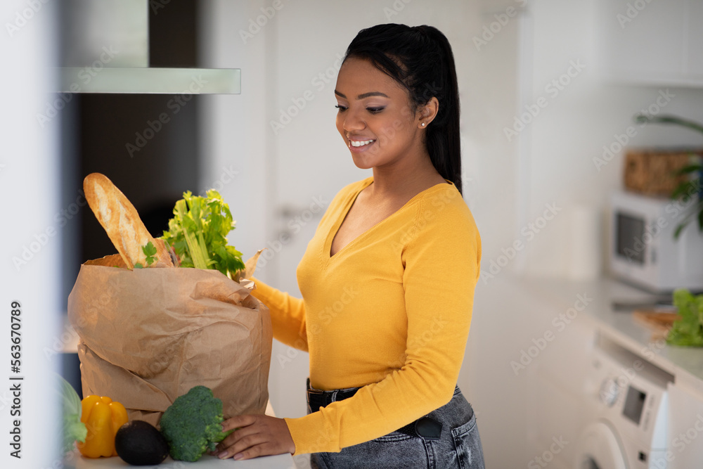 Wall mural Smiling young african american woman in casual unpacks paper bag with groceries and organic vegetables