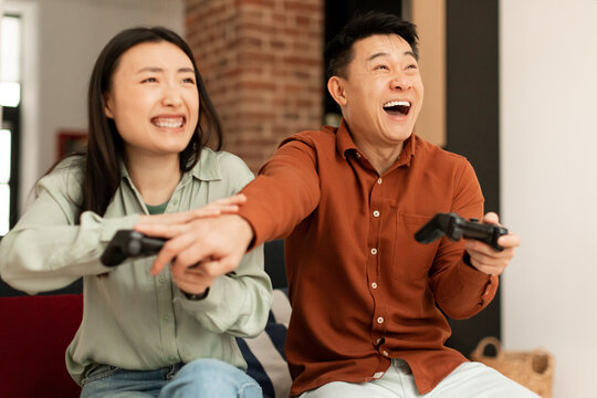 Excited Asian Couple Playing Video Games At Home, Happy Asian Emotional Man And Woman Sitting On Couch With Joysticks