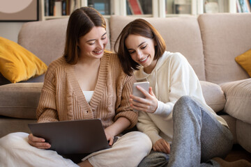 Smiling european young women with laptop and smartphone surfing in internet, chatting, show video