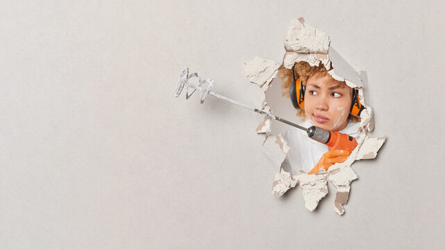 Serious female builder does renovation in her house holds paint mixer in hands breaks through white wall uses headphones for noise cancellation focused aside. Redecoration and repair concept.