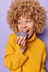 Curly haired woman bites delicious glazed doughnut looks aside has sweet tooth wears yellow jumper...