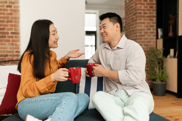 Happy asian spouses sitting on sofa with cups of hot drink and talking, resting in living room interior