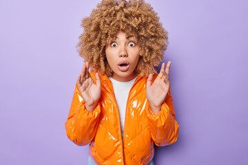 Studio shot of scared young woman raises hands up being startled keeps mouth opened wears orange jacket reacts to something impressive isolated over purple background. Human reactions concept