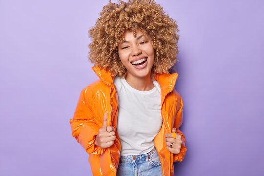 Happy cheerful woman laughs joyfully expresses positive emotions smiles broadly shows white teeth wears white basic t shirt jeans and orange jacket isolated over purple background. Happiness concept