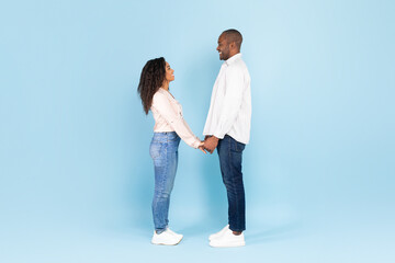 Full length of black couple holding hands and looking at each other against blue background, side view, free space