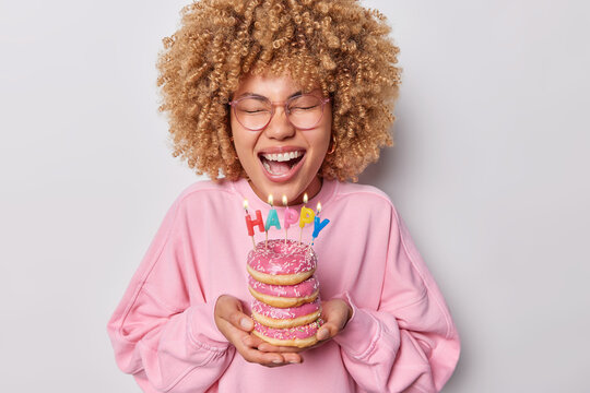 Positive curly haired woman wears spectacles and pullover holds pile of delicious doughnuts with letter shaped candles enjoys festive occasion laughs out gladfully isolated over grey background