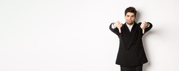 Portrait of skeptical and disappointed man in black suit, frowning upset, showing thumbs-down,...