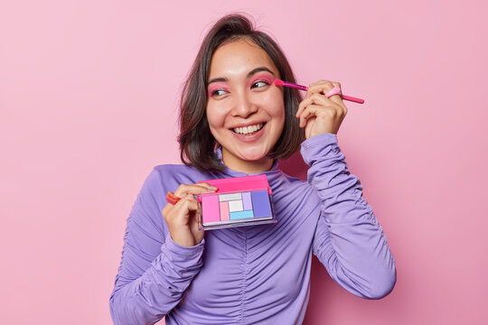 Happy Asian woman with dark hair applies pink eyeshadow uses cosmetic brush smiles broadly has white even teeth wears purple turtleneck isolated over pink background. Beauty and wellness concept