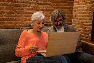 elderly married couple, home retirement camera sitting next to laptop with credit card. Satisfied senior spouse digital bank customers paying online purchases - focus in woman -