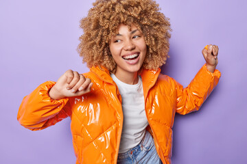 Curly haired woman smiles broadly shows white teeth dances carefree has playful expression wears fashionable orange jacket has happy mood isolated over purple background. Cheerful female dancer