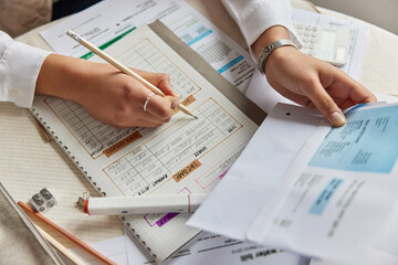 Fototapeta premium Money and finances. Unrecognizable woman tries to find way to solve financial problems holds bill writes down notes in notebook plans expenses holds pencil prepares business report. View from above