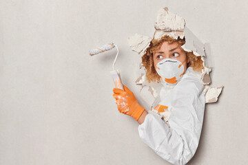 Professional female architect builder holds paint roller wears protective respirator and costume works on interior design and renovation breaks through plaster wall. Painter in uniform with tools