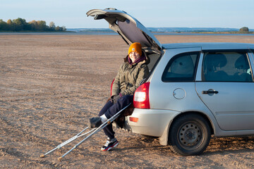 A young girl sits on a car trunk with a broken leg, on crutches, admiring the sunset on a sandy...